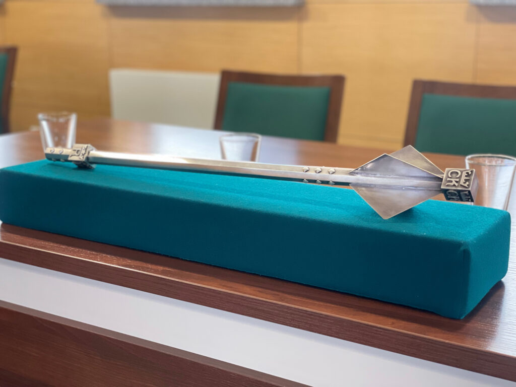 Srebrny berło z symbolem CK na zielonej poduszce. Kontekst: sala konferencyjna, stół, krzesła.