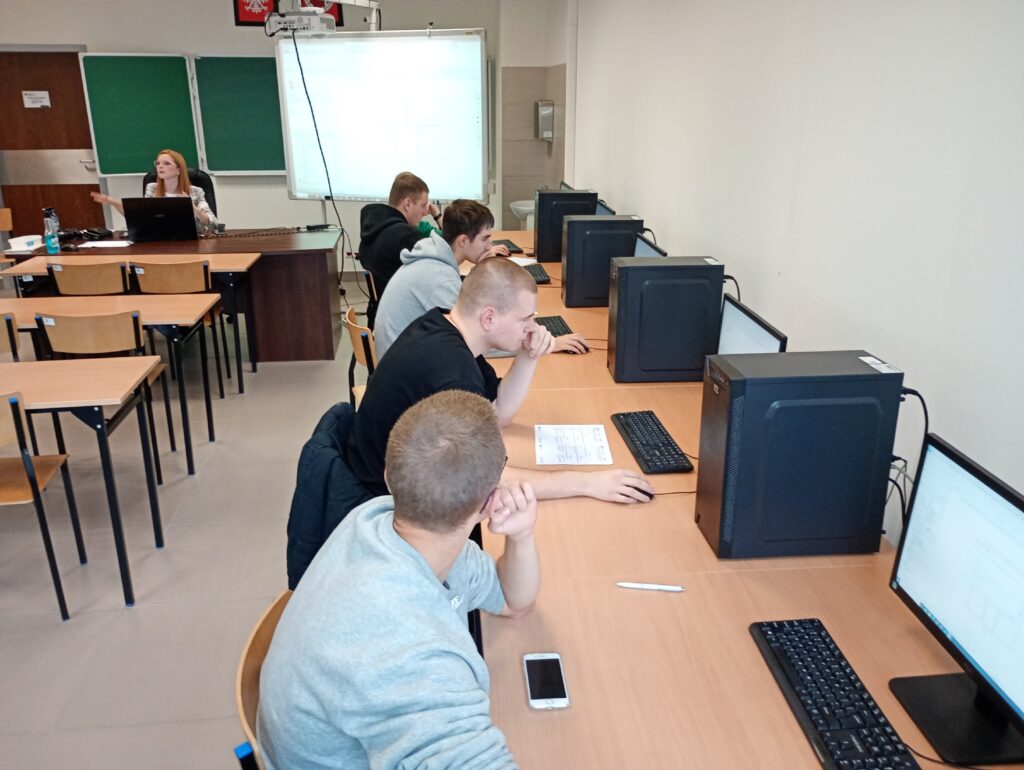 Sala komputerowa z uczniami przy stanowiskach. Kobieta przy biurku z laptopem.