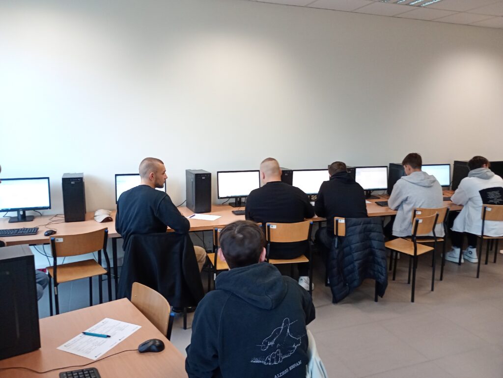 Grupa osób siedzi przy komputerach w sali lekcyjnej. Kluczowe obiekty: komputery, krzesła, biurka. Kontekst: sala komputerowa.