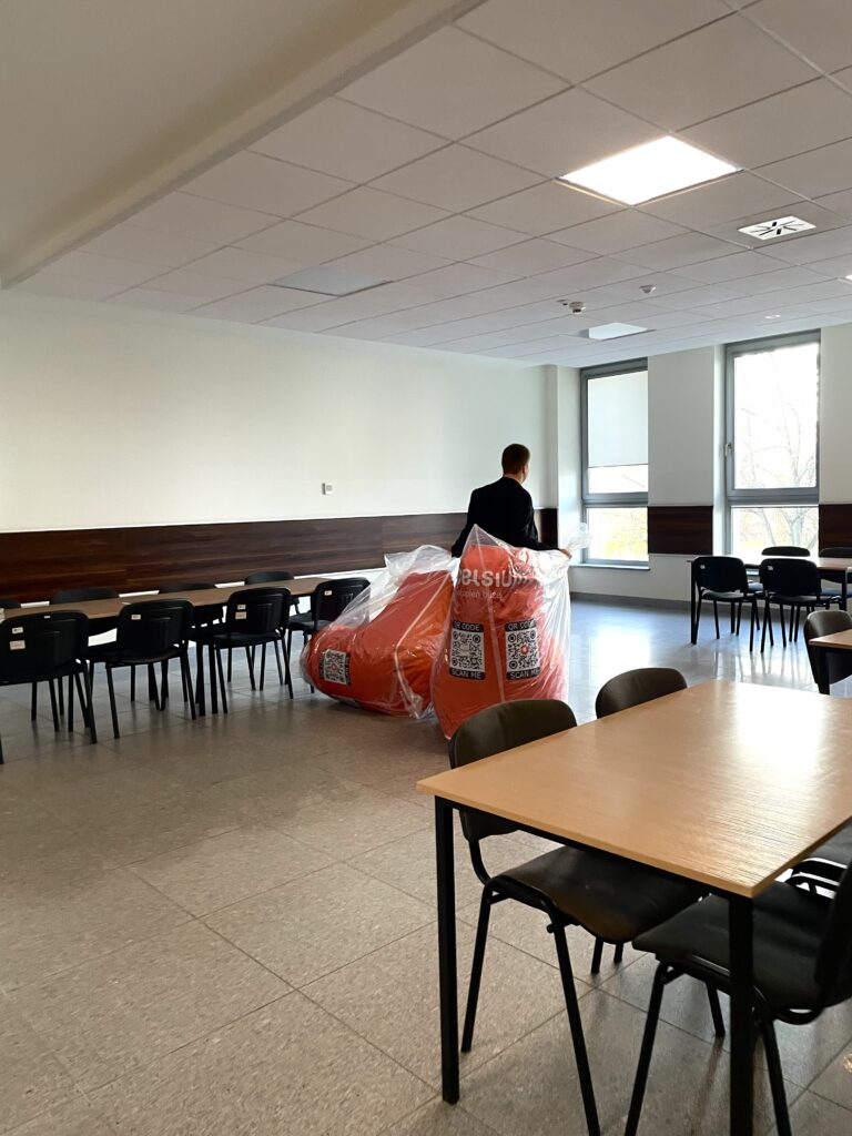 Osoba w czarnym ubraniu przenosi dwa duże, czerwone worki w przezroczystej folii.