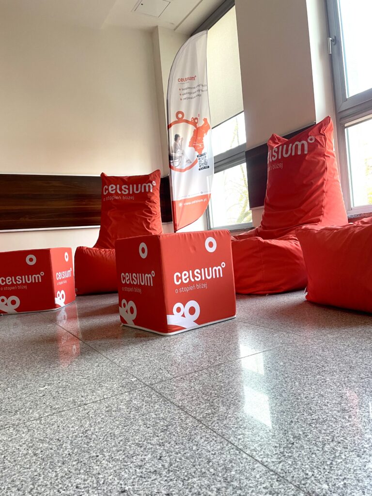 Czerwone pufy i kostki z logo "celsius" stoją na szarym, polerowanym podłożu. W tle biała flaga reklamowa.