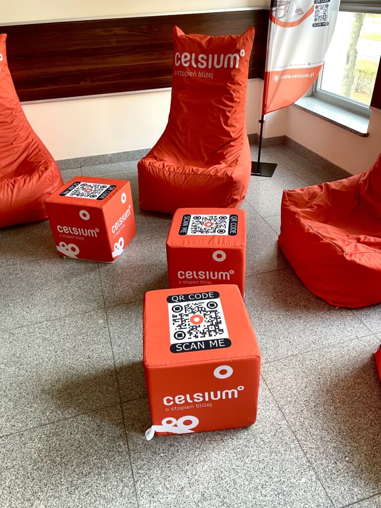 Pomarańczowe pufy i kostki z logo "celsius" i kodami QR. Kontekst: przestrzeń eventowa lub poczekalnia.