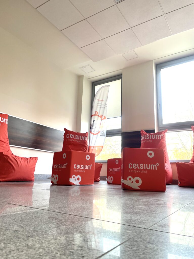 Czerwone pufy i kostki z logo "celsium" stoją na błyszczącej podłodze. W tle okna i baner.