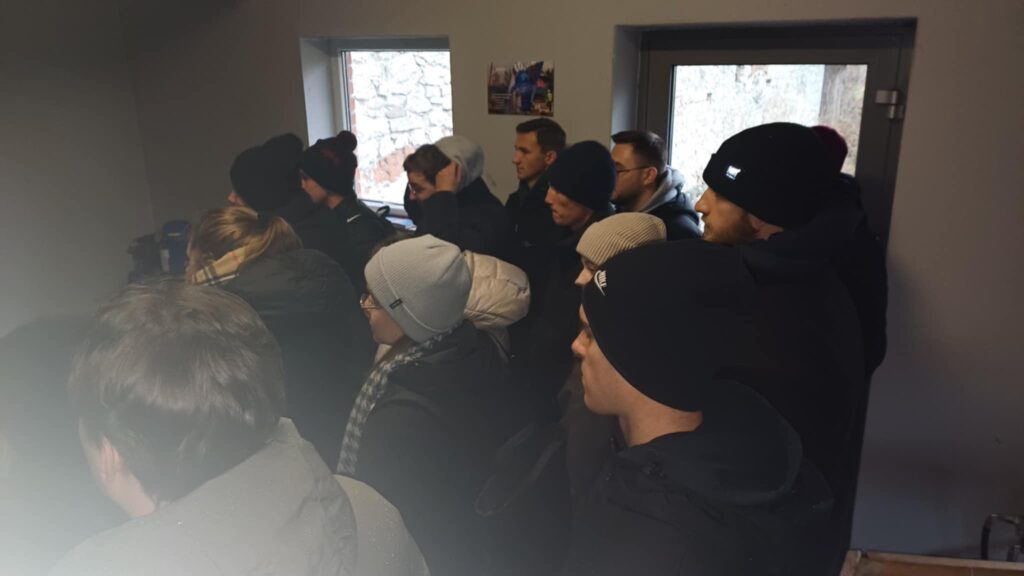Grupa osób w zimowych ubraniach stoi w pomieszczeniu. Kluczowe obiekty: czapki, kurtki, okno.