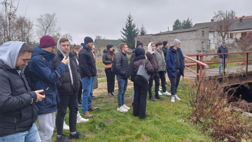 Grupa ludzi stoi na trawie. W tle budynki i most.