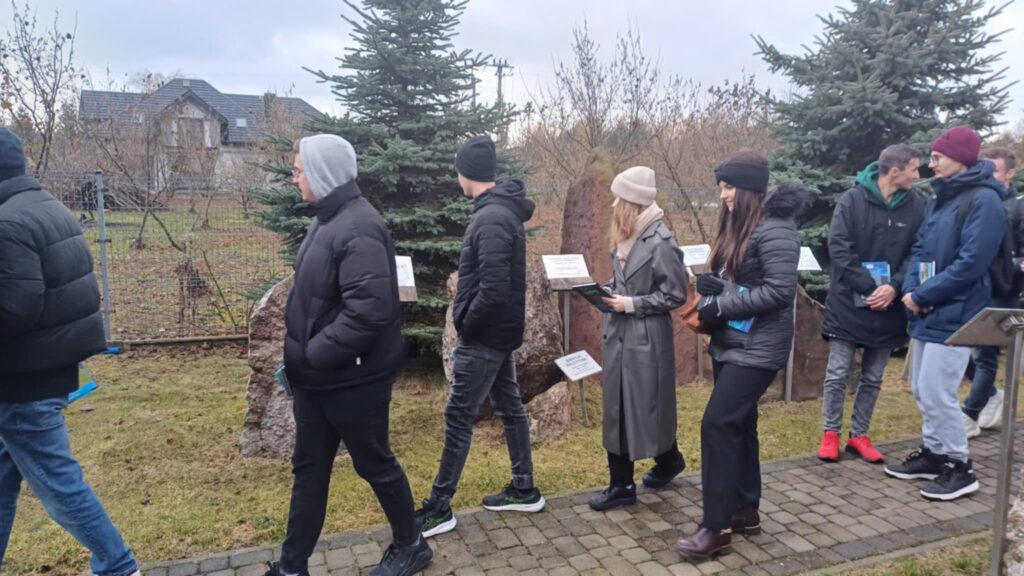 Grupa osób ogląda kamienie z tablicami informacyjnymi. W tle dom i drzewa.