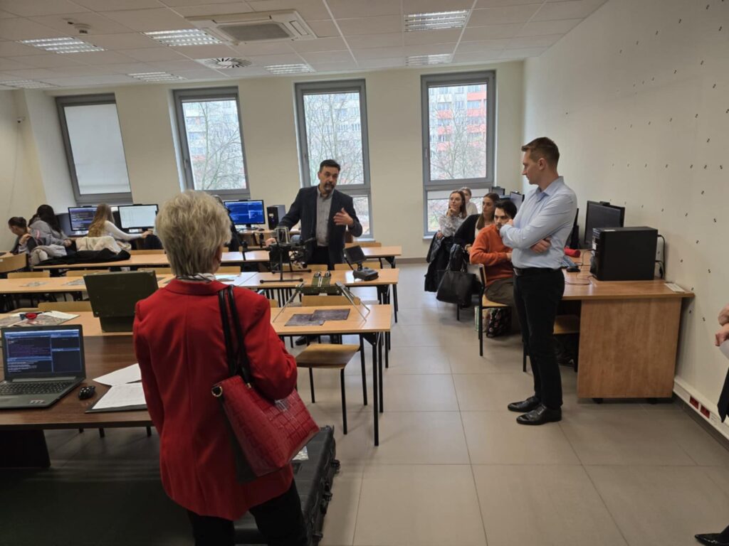 Sala lekcyjna z komputerami i ludźmi. W centrum mężczyzna w garniturze prezentuje sprzęt.