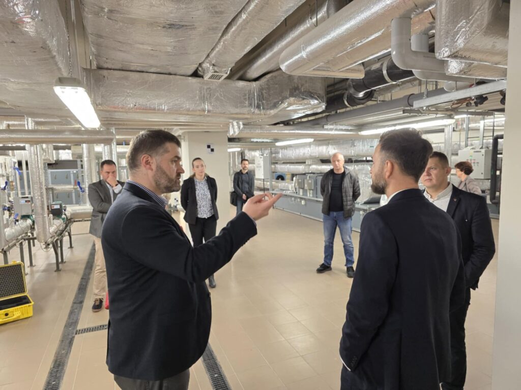 Grupa osób w pomieszczeniu technicznym z rurami i izolacją. W tle widoczne są urządzenia.
