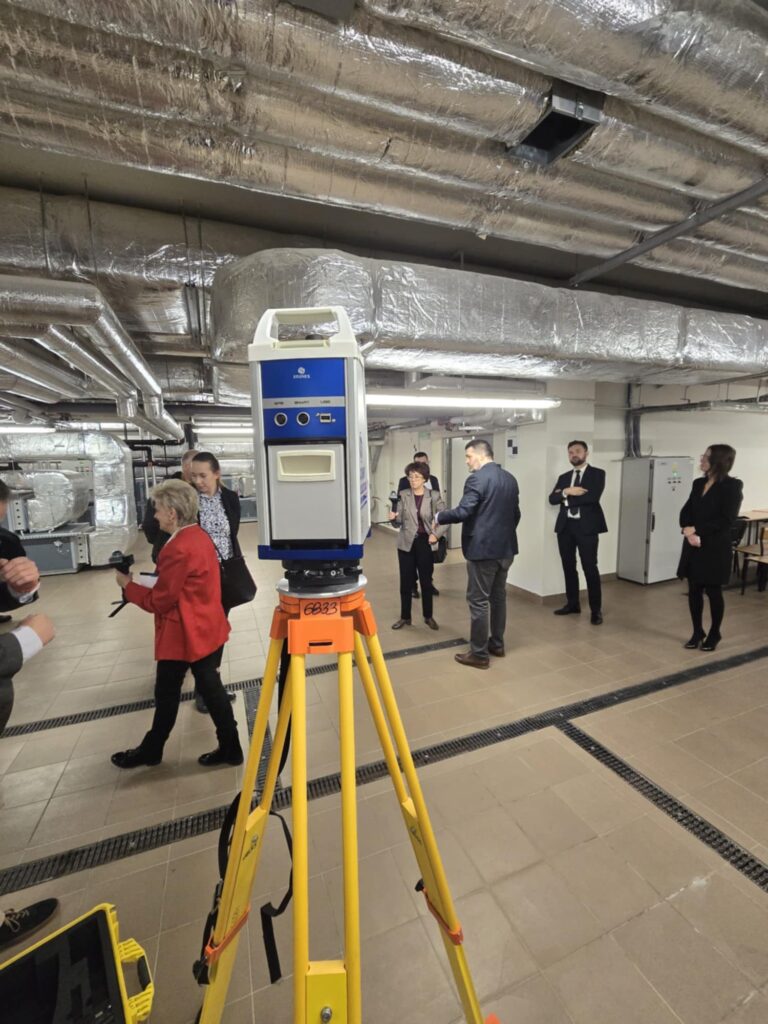 Grupa osób obserwuje skaner 3D na statywie w pomieszczeniu technicznym z widocznymi instalacjami.