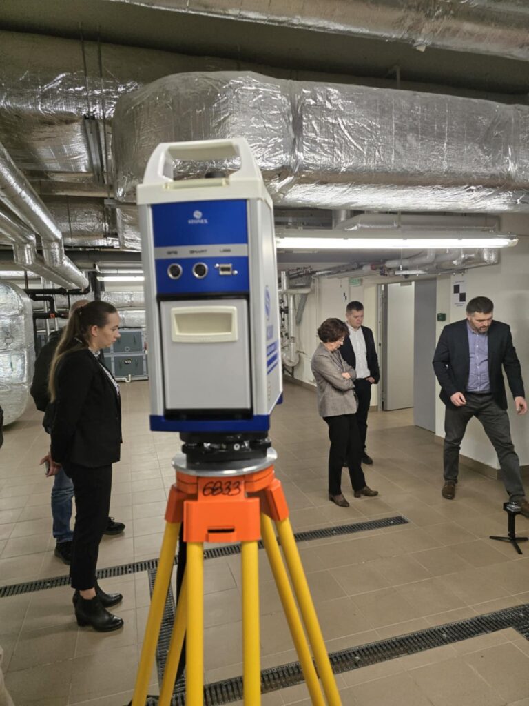 Skaner 3D na statywie w pomieszczeniu technicznym z rurami i ludźmi. Kluczowe obiekty: skaner 3D, statyw, rury, ludzie.