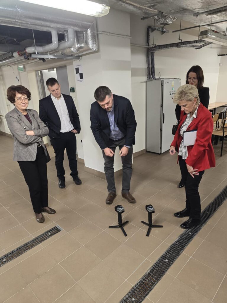 Osoby w pomieszczeniu z rurami i szafami elektrycznymi oglądają dwa urządzenia na statywach.
