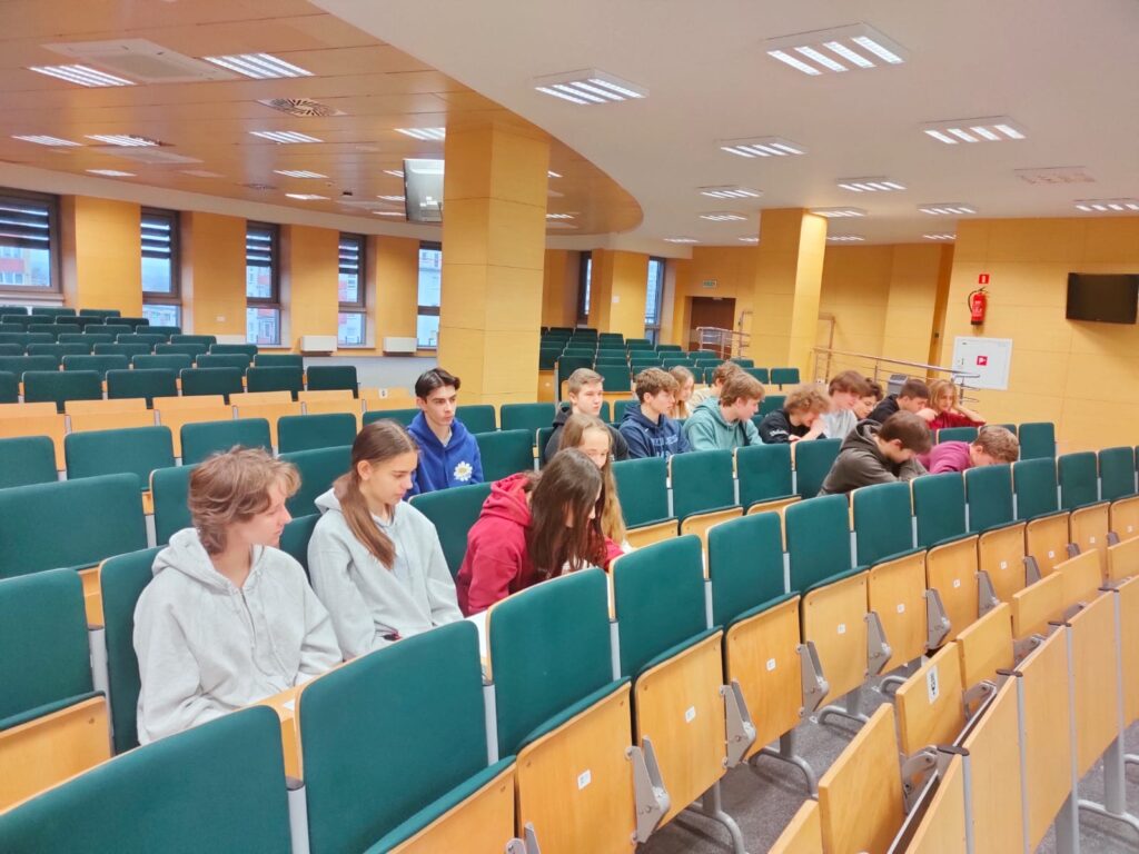 Grupa młodzieży siedzi w sali wykładowej. Widoczne są rzędy zielonych siedzeń i drewniane stoły.