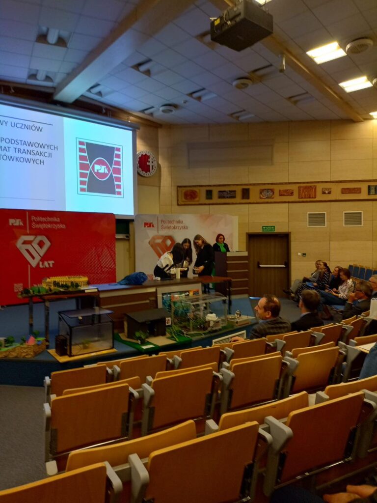 Sala wykładowa z widownią, ekranem projekcyjnym i wystawą modeli. Kontekst: wydarzenie edukacyjne lub prezentacja.