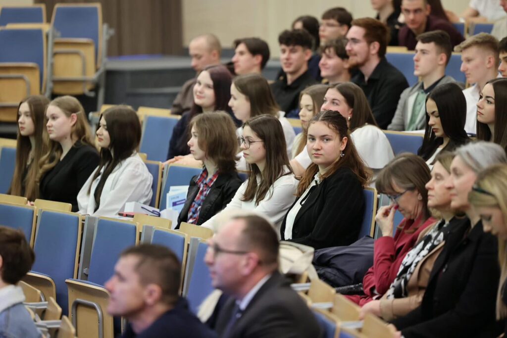 Grupa ludzi siedzi na niebieskich krzesłach w sali wykładowej. Widać studentów i wykładowców.