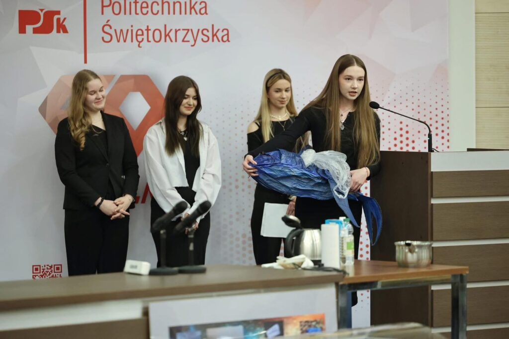 Cztery młode kobiety stoją przed ścianą z logo Politechniki Świętokrzyskiej. Jedna z nich trzyma niebieski przedmiot.