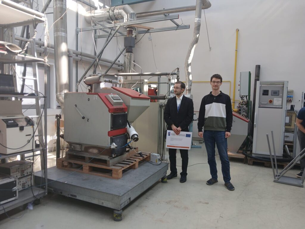 Dwóch mężczyzn stoi obok urządzenia w laboratorium. Kluczowe obiekty: urządzenie, rury, mężczyźni.