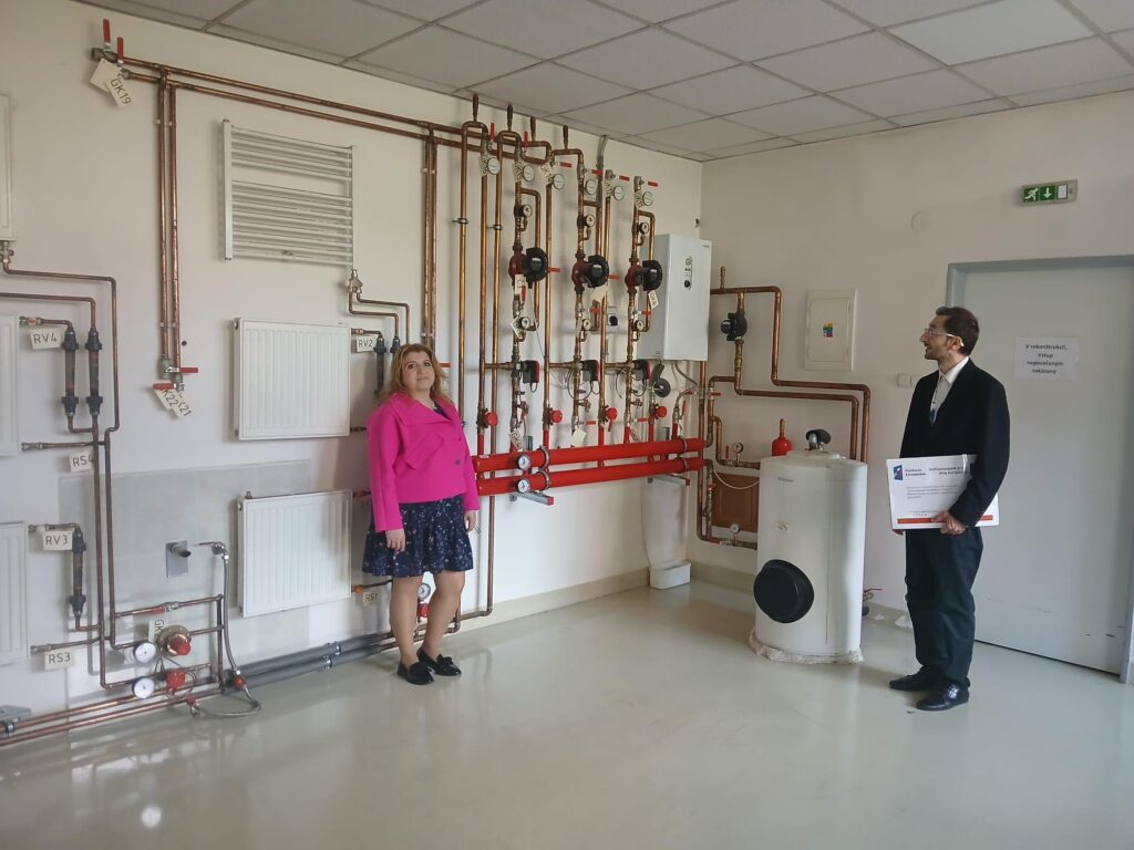 Dwie osoby stoją przy instalacji grzewczej z miedzianymi rurami, grzejnikami i bojlerem.