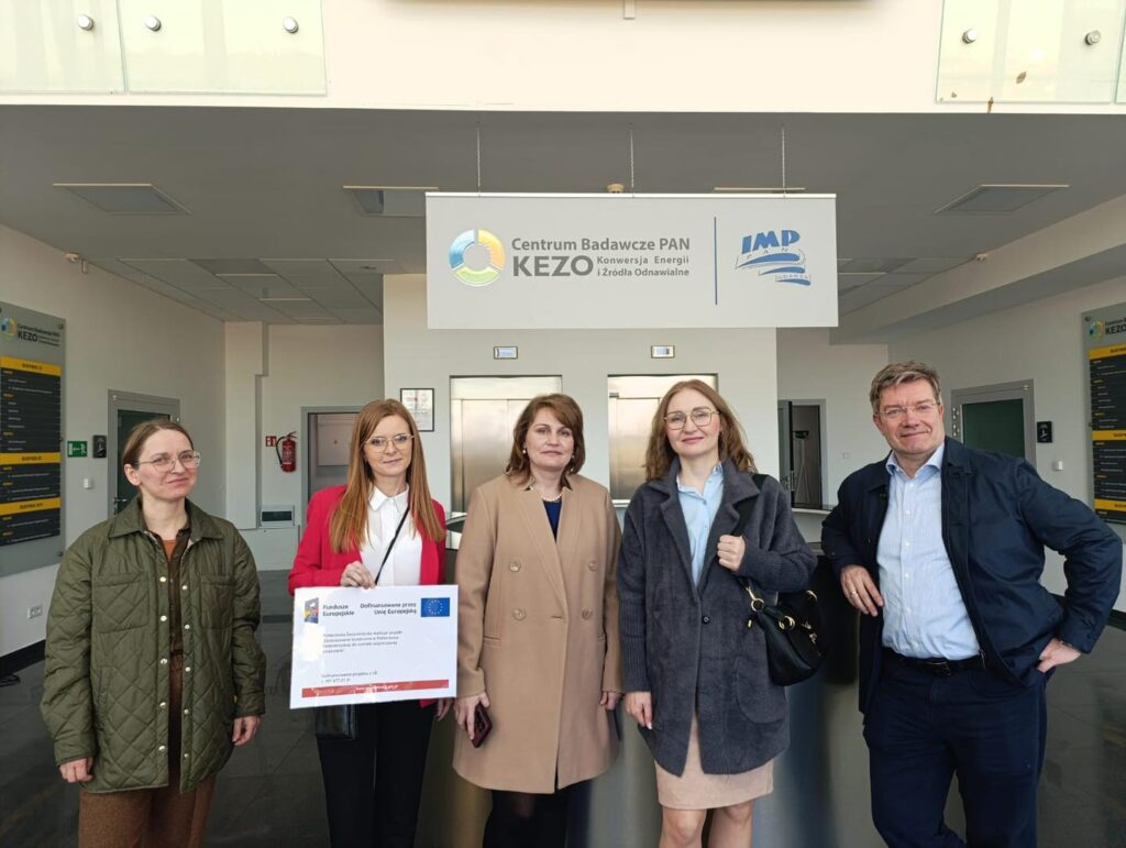 Cztery osoby stoją przed banerem Centrum Badawcze PAN KEZO. Jedna osoba trzyma dokument z logiem UE.