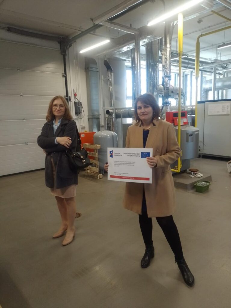 Dwie kobiety stoją w pomieszczeniu z rurami i urządzeniami. Jedna trzyma tablicę z logo UE.