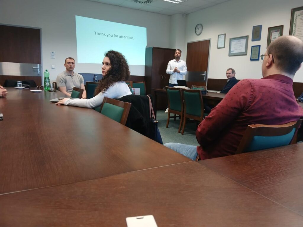 Ludzie siedzą przy stole konferencyjnym. Na ekranie widnieje napis "Thank you for attention". Kontekst: spotkanie.