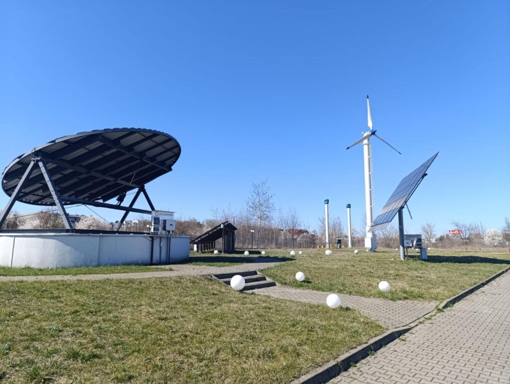 Duży panel słoneczny, turbina wiatrowa i mniejsze panele słoneczne na trawiastym terenie.