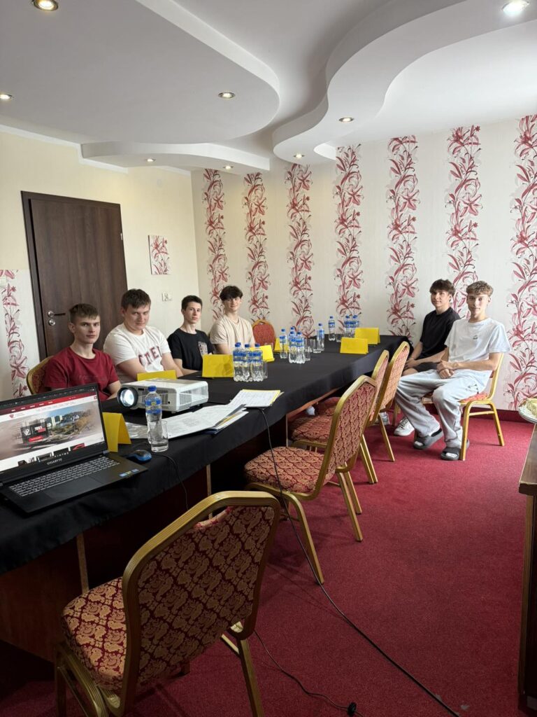 Grupa młodych mężczyzn siedzi przy stole konferencyjnym. Na stole laptop, projektor i butelki z wodą.