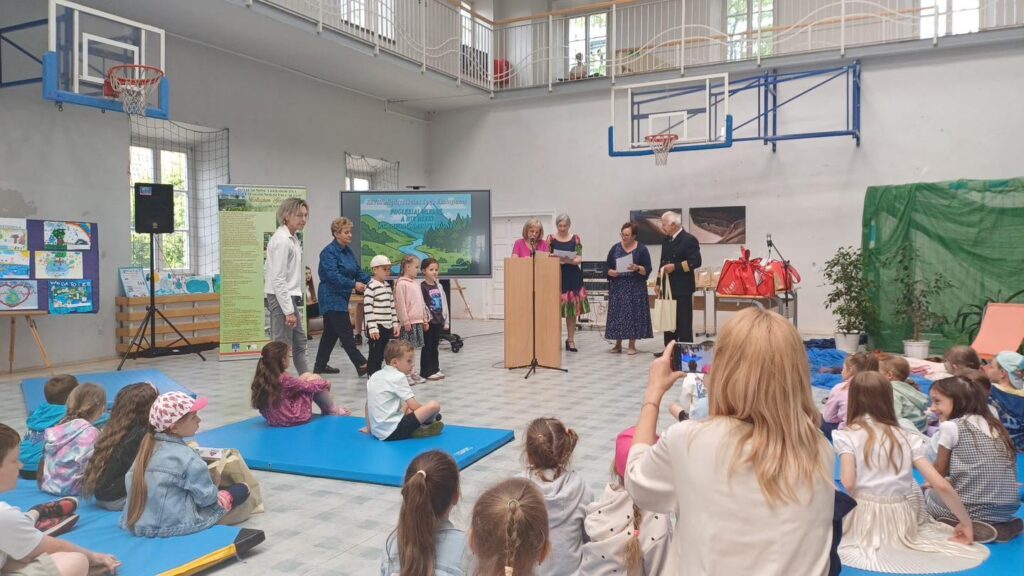 Grupa dzieci i dorosłych w sali gimnastycznej. Na ekranie widnieje napis "POGŁĘBIALNIA WODY".