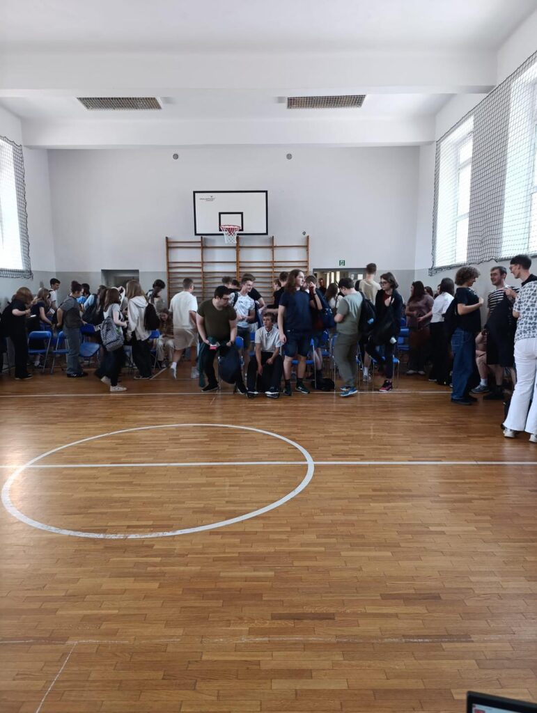 Grupa ludzi stoi na sali gimnastycznej. Widać kosz do koszykówki, drewnianą podłogę i siatkę.