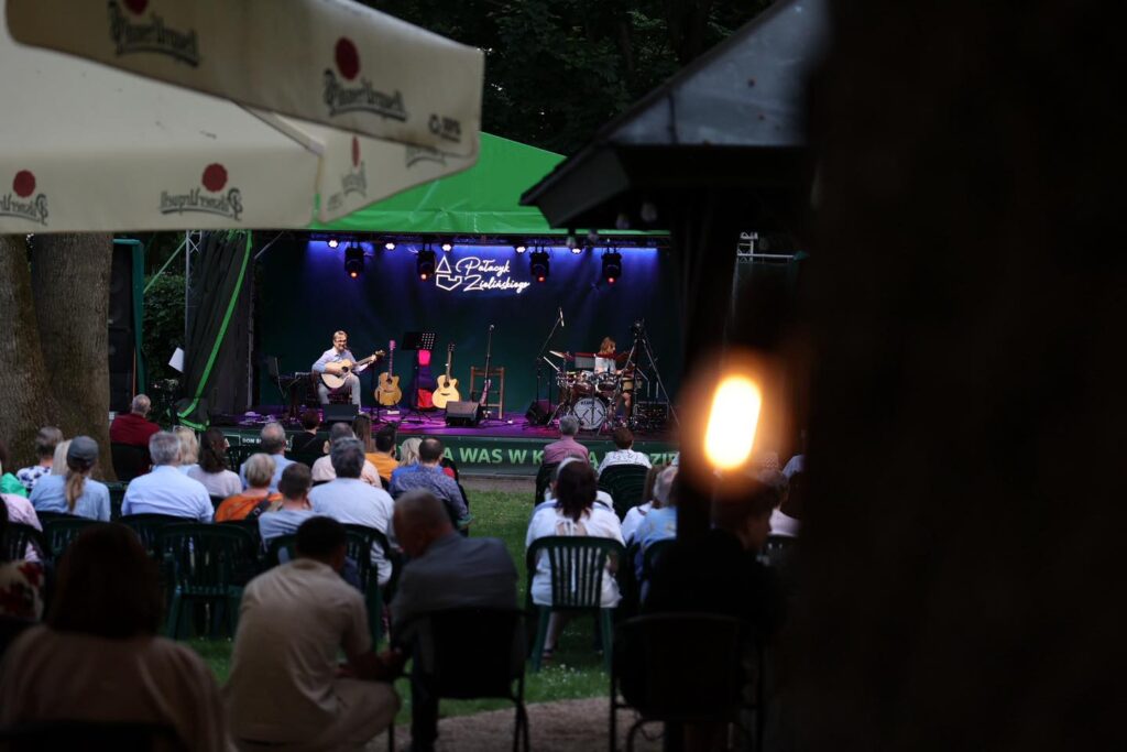 Koncert na świeżym powietrzu. Muzycy grają na scenie, publiczność siedzi na krzesłach.