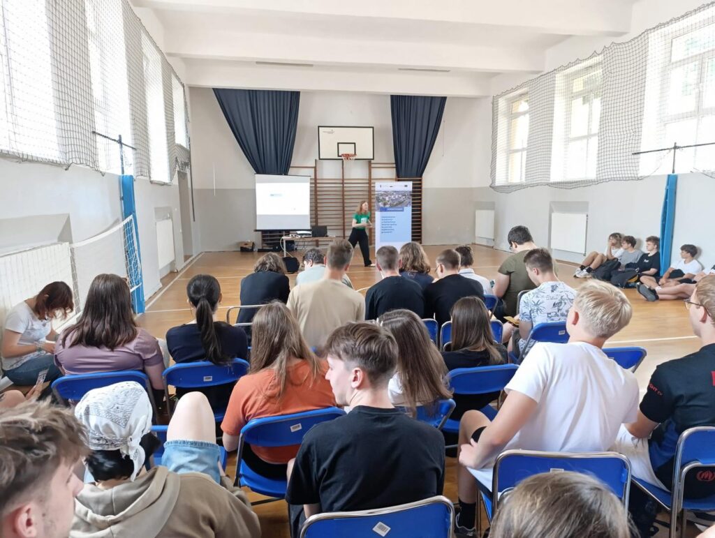 Sala gimnastyczna z widownią siedzącą na krzesłach. Na ekranie wyświetlana jest prezentacja.
