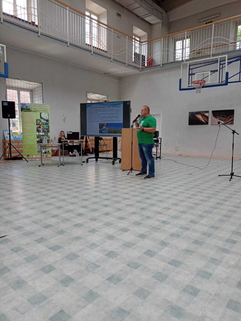 Mężczyzna przemawia na tle ekranu z prezentacją. W sali znajdują się też inne osoby i plakaty.