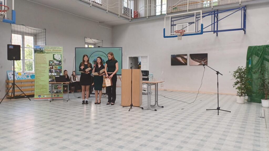 Trzy młode kobiety stoją przy podium w sali gimnastycznej. Kluczowe obiekty: podium, mikrofon, baner, kosz do koszykówki.