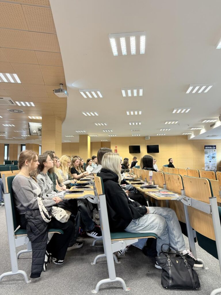 Grupa studentów siedzi w sali wykładowej. Na ścianie wisi plakat.