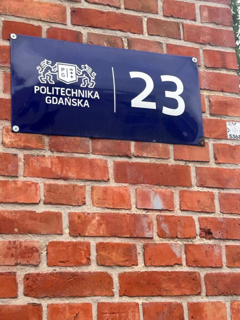 Niebieska tablica z logo Politechniki Gdańskiej i numerem 23, zamontowana na ceglanej ścianie.