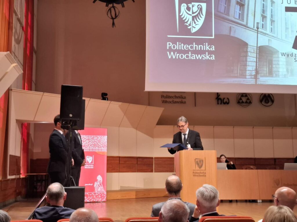 Mężczyzna w garniturze przemawia przy mównicy Politechniki Wrocławskiej. W tle widownia i logo uczelni.
