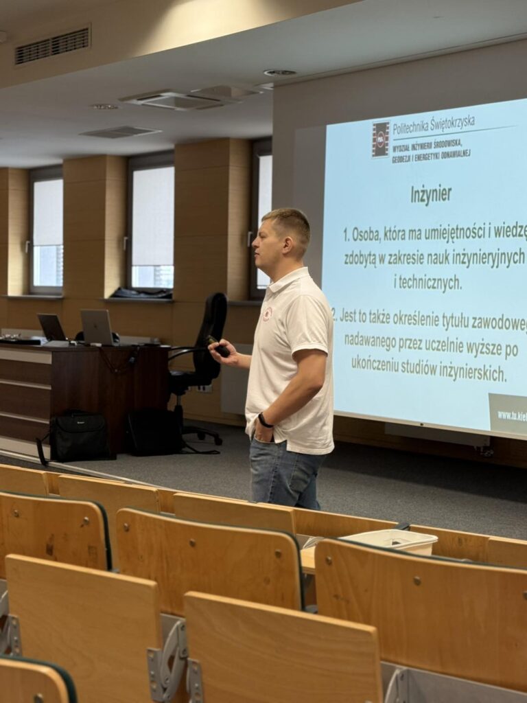 Mężczyzna w białej koszulce prezentuje definicję inżyniera na ekranie. W tle sale wykładowe.