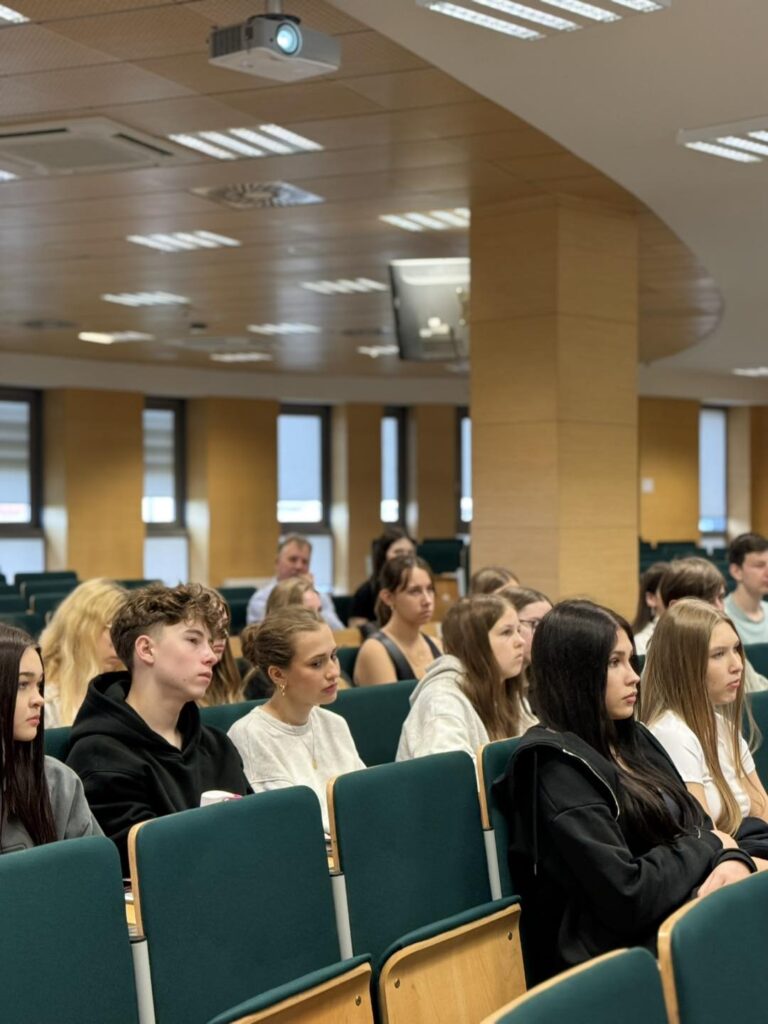 Grupa młodych ludzi siedzi w sali wykładowej. Widoczne są: projektor, krzesła, ludzie.