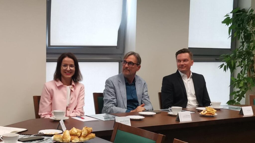 Trzy osoby siedzą przy stole, na którym są filiżanki i ciastka.