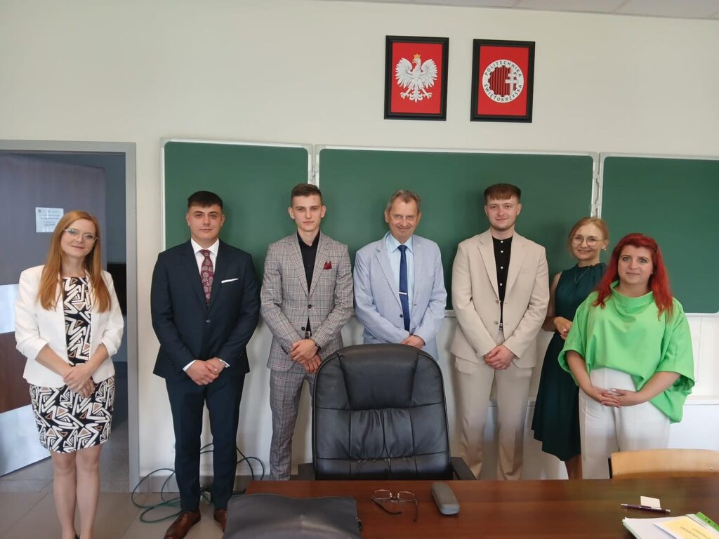 Grupa osób stoi przed zieloną tablicą. W tle widnieją godła.
