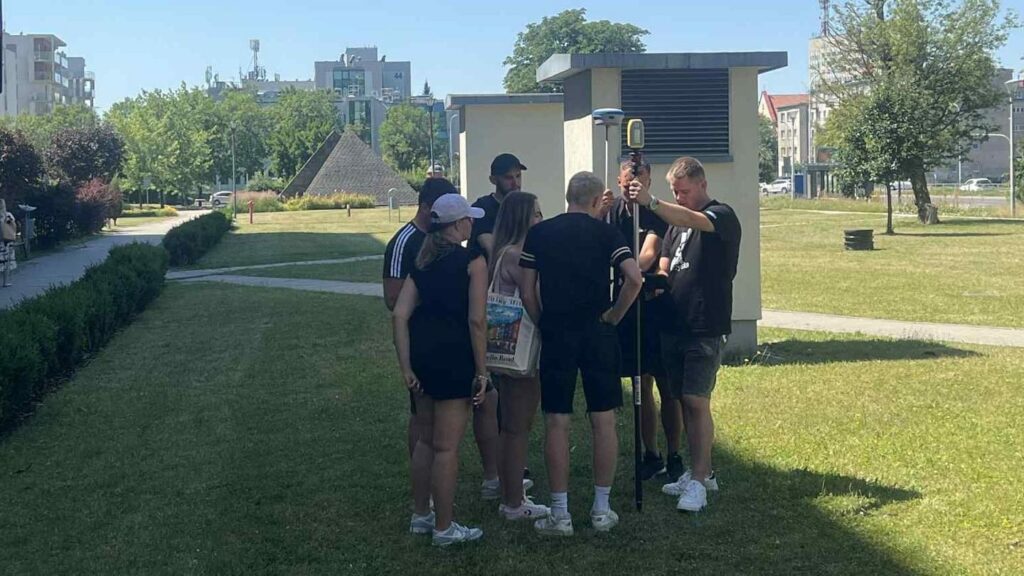 Grupa osób stoi wokół geodezyjnego sprzętu pomiarowego na trawiastym terenie, w tle budynki i drzewa.