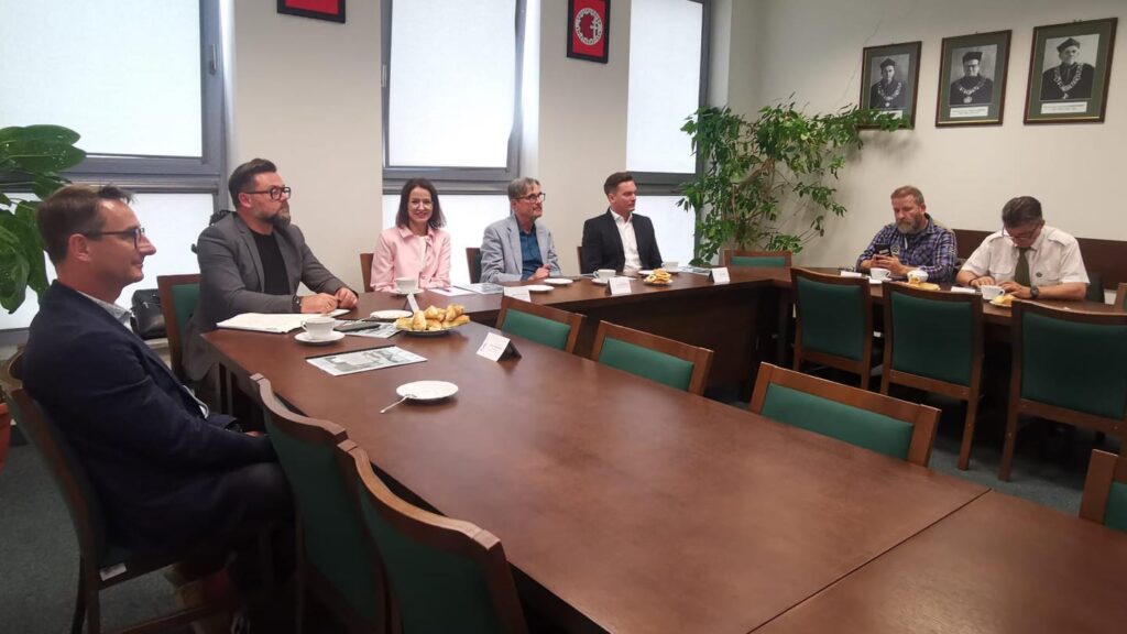 Grupa osób siedzi przy stole konferencyjnym. Widać dokumenty, filiżanki i talerze z przekąskami.