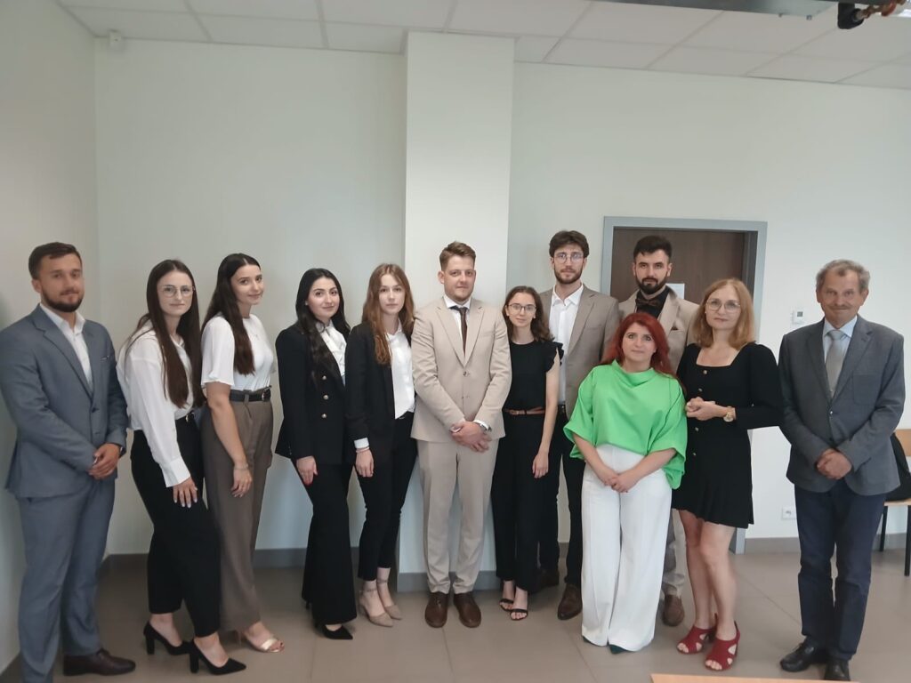Grupa osób w strojach formalnych stoi w pomieszczeniu. Kluczowe obiekty: ludzie, garnitury, sukienki.