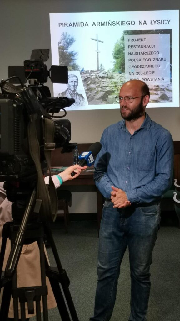 Prezentacja o Piramidzie Armińskiego na Łysicy. Kluczowe obiekty: ekran z napisem, mężczyzna, kamera.