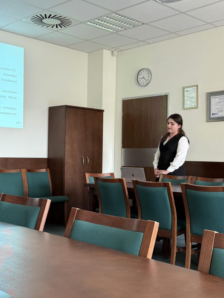 Kobieta w sali konferencyjnej przy stole z laptopem. W tle szafa, zegar, certyfikaty.