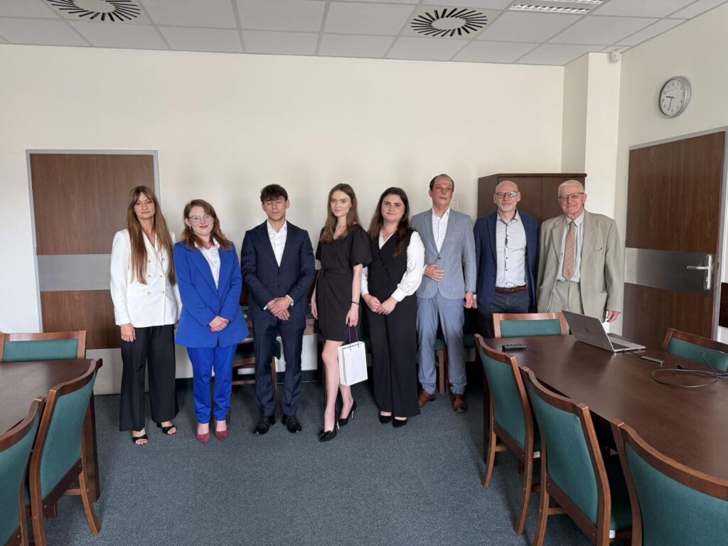 Osiem osób w formalnych strojach stoi w sali konferencyjnej. W tle drzwi, szafa, zegar i stół z laptopem.