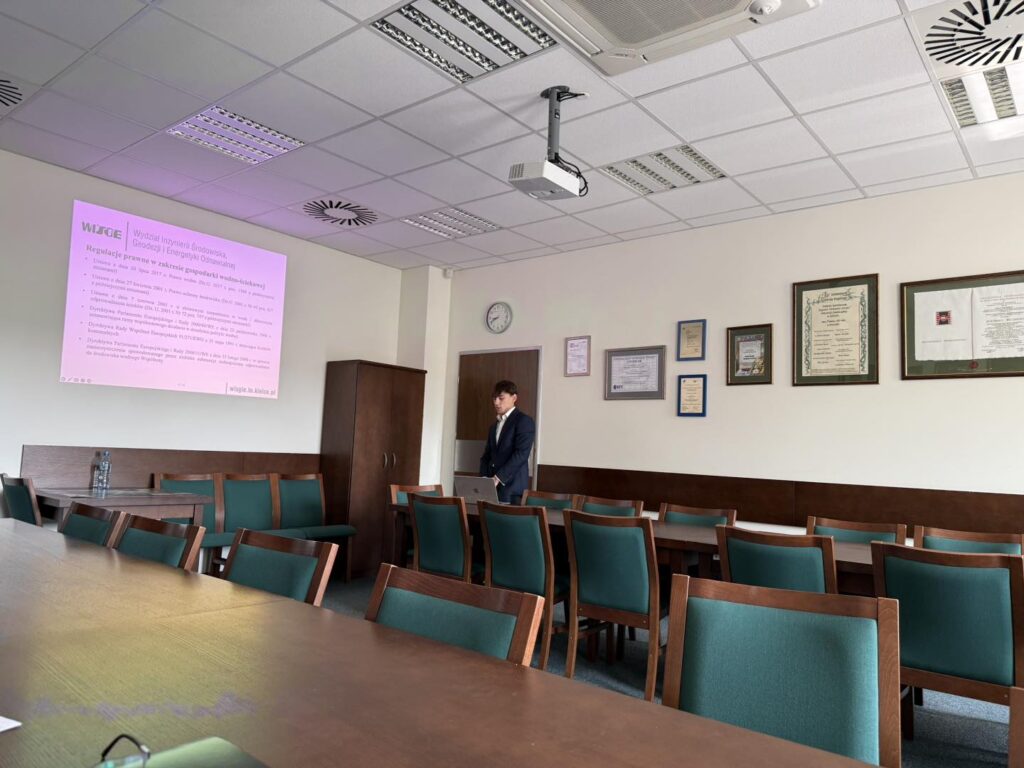 Sala konferencyjna z projektorem, ekranem, stołami i krzesłami. Na ścianie wiszą dyplomy.