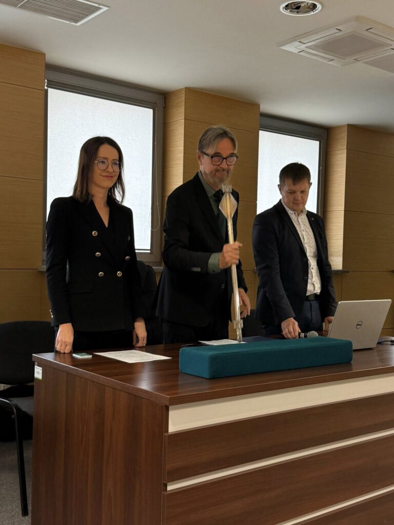 Trzy osoby stoją przy biurku. Mężczyzna trzyma berło. Na biurku laptop i mikrofon.