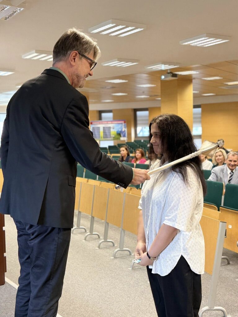 Mężczyzna w garniturze podaje kobiecie laskę. Kontekst: sala wykładowa z widownią.