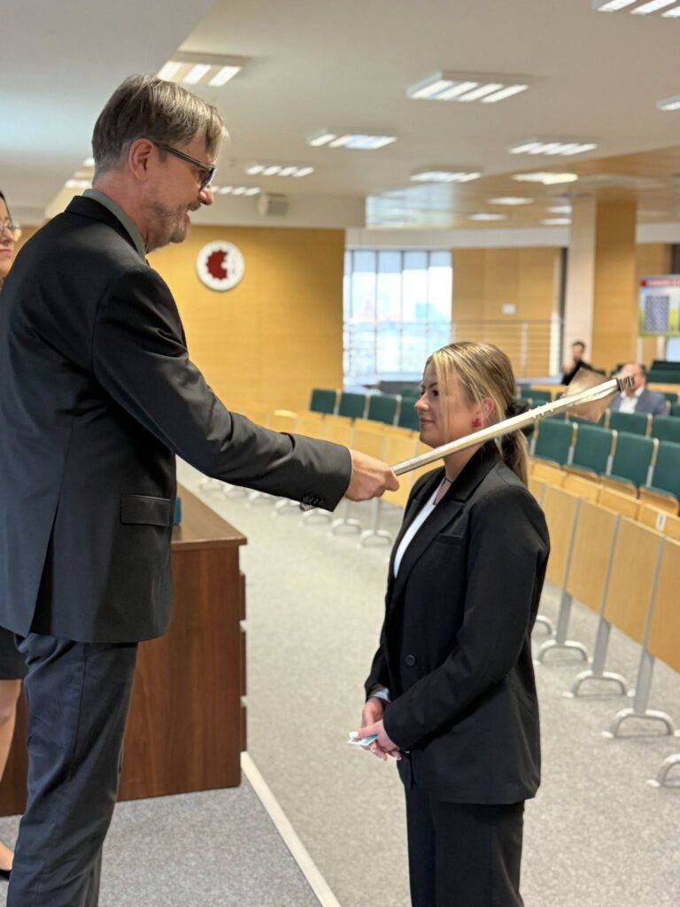 Mężczyzna w garniturze podaje kobiecie w garniturze laskę. Kontekst: sala wykładowa.