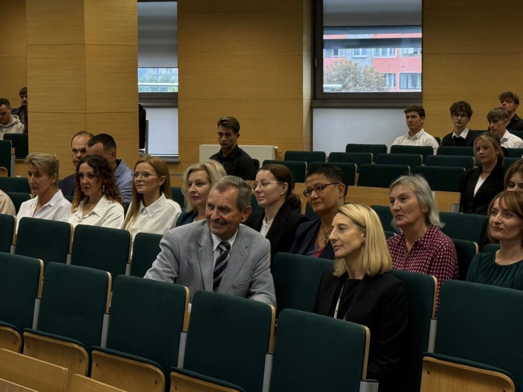 Ludzie siedzą w sali wykładowej. Widać krzesła, drewniane panele i okna.